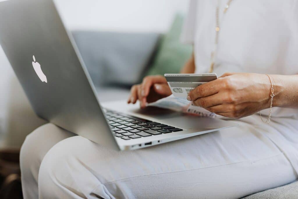 Persona usando una laptop MacBook mientras sostiene una tarjeta de crédito, realizando una compra o transacción en línea desde el sofá.
